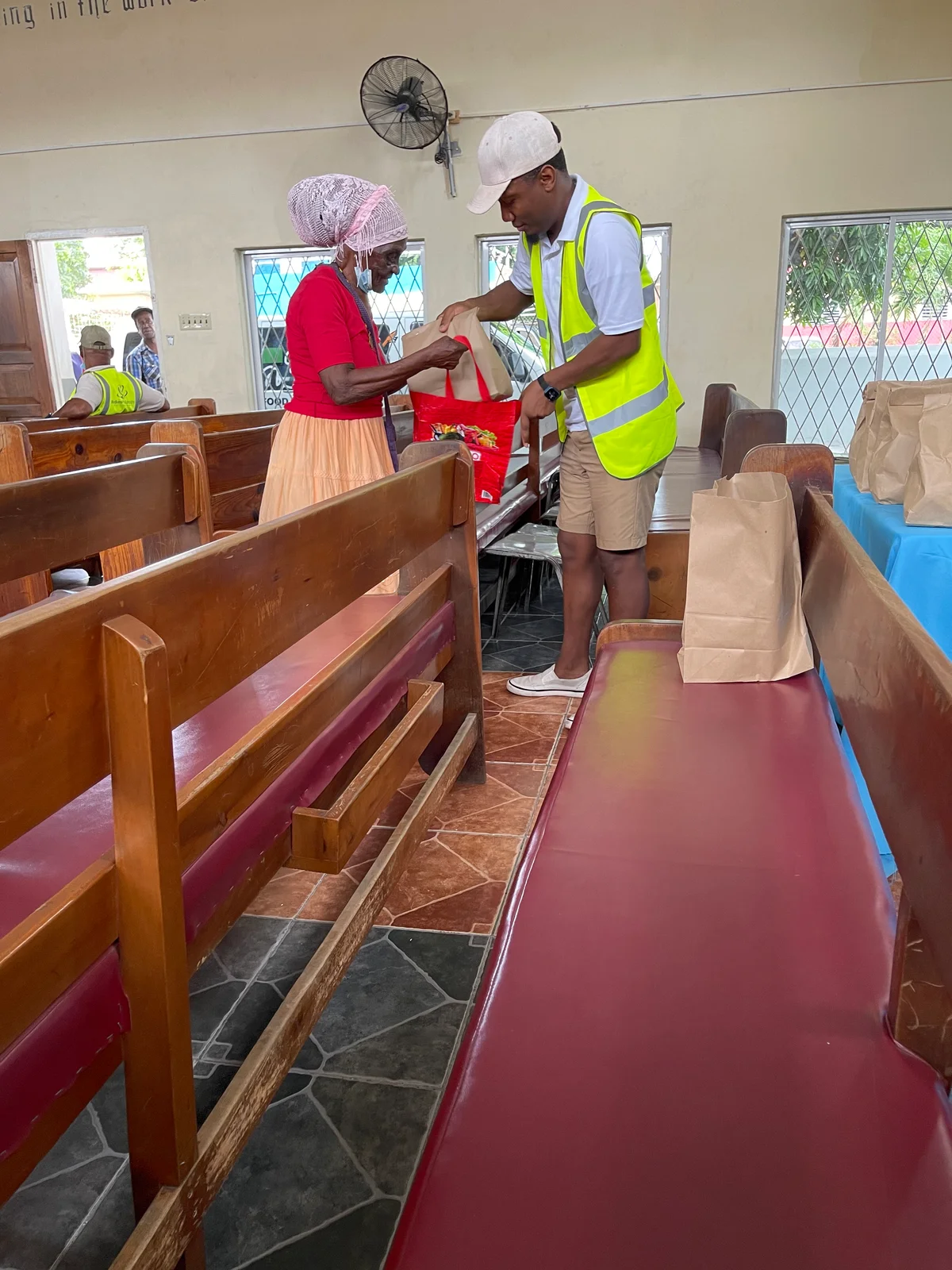 People near a table and bags in a hall with wooden benches.